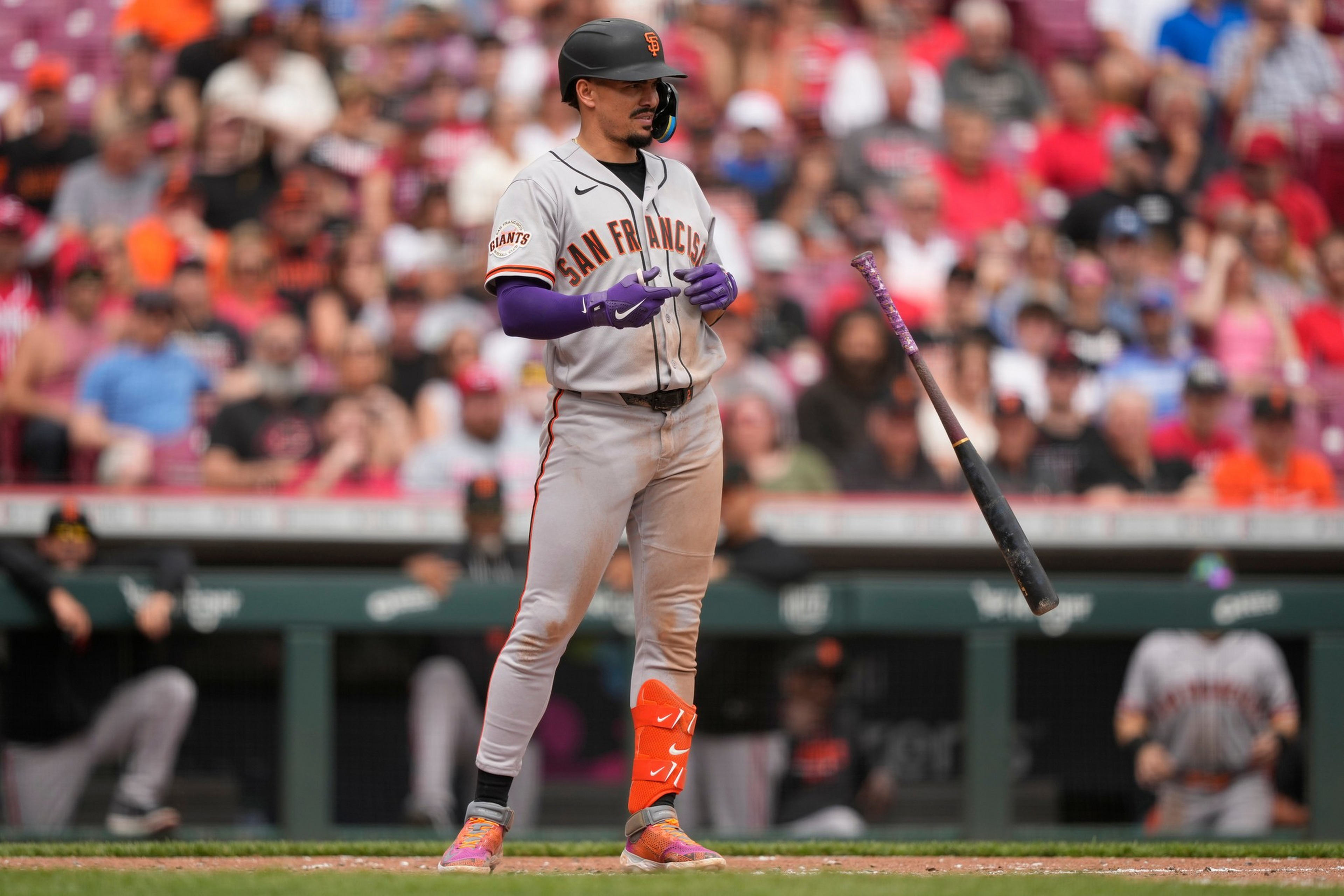 A San Francisco Giants player stands at bat, wearing gray and orange gear, as his bat floats midair after being tossed up.