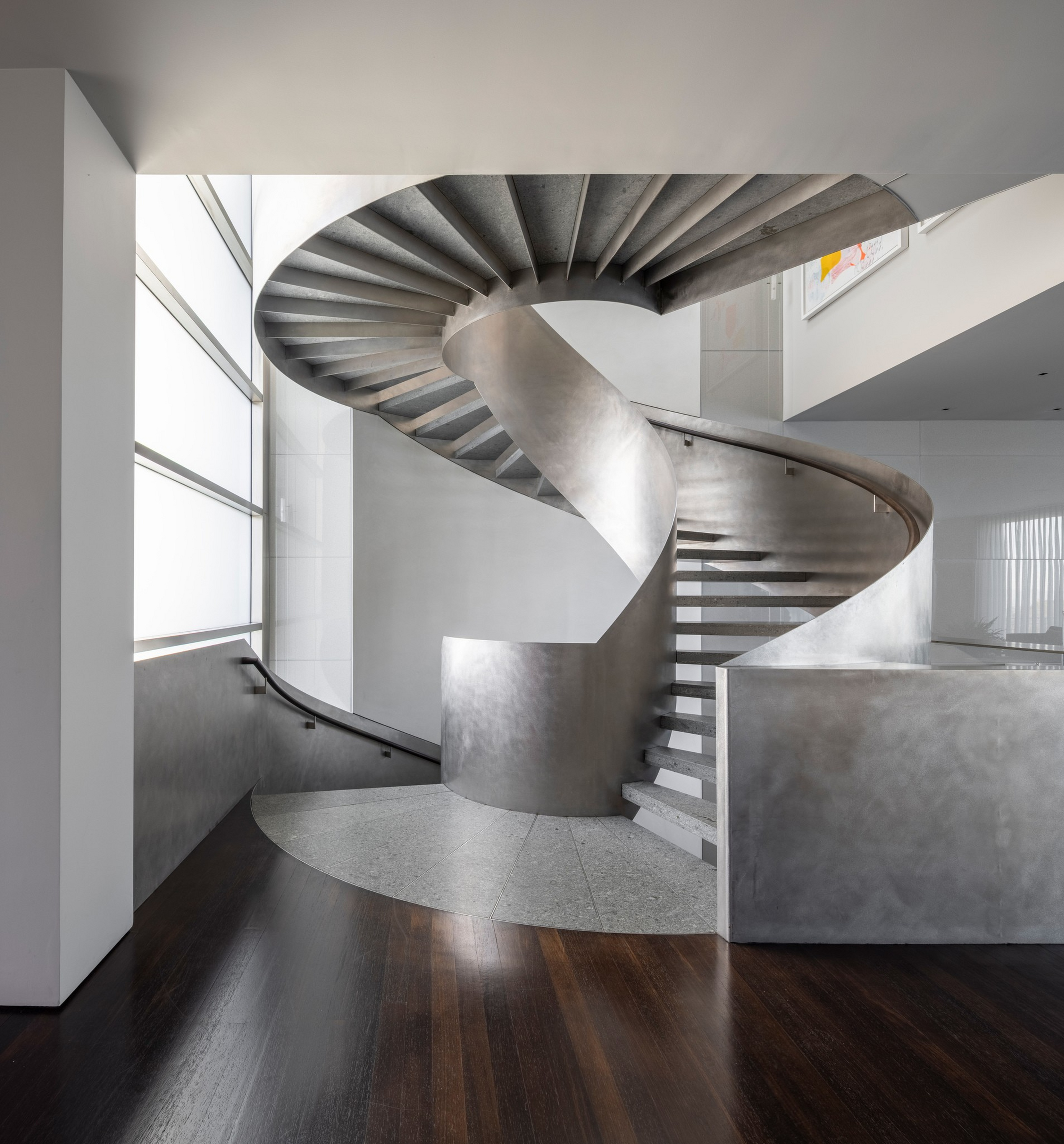 A sleek, modern spiral staircase made of smooth metal curves upward in a bright, minimalist space with wooden and terrazzo flooring.
