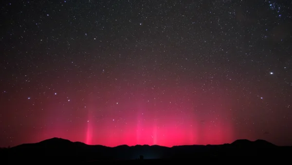 Rippling red light emerges into a starry night sky from behind a hilly silhouette.