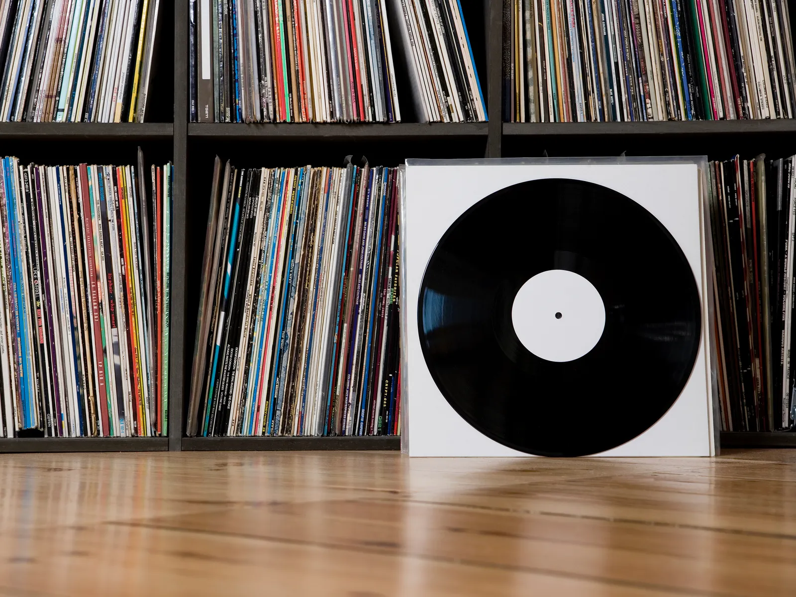 A vinyl record in a white protective sleeve leaning up against shelves full of more records