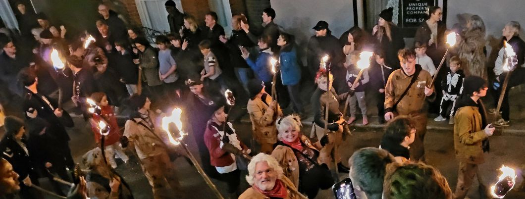 A shot of a section of the Hastings Borough Bonfire Society’s torchlit procession showing people carrying burning torches down a narrow street with onlookers on either side of them