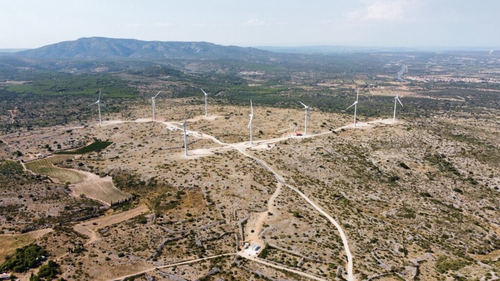 Eight wind turbines top an arid hill, with greener mountains in the distance.